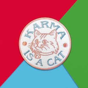 Könnte beinhalten: Runder, gestickter Aufnäher mit einer Katzenillustration und dem Text "Karma is a Cat" in Blau. Der Aufnäher hat einen weißen Hintergrund und einen rosa Rand. Der Hintergrund ist in rote, blaue und grüne Abschnitte unterteilt.