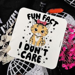 Könnte beinhalten: Ein quadratischer weißer Untersetzer mit einem Cartoon-Kätzchen in Orange, einer rosa Schleife und einem Halsband mit Glöckchen. Der Text FUN FACT I DON'T CARE ist von schwarzen Sternen umgeben. Rechts steht der kleine Text GRINDSTORE LTD.