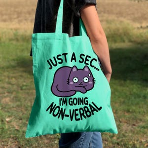 Op de afbeelding: Een turquoise tote bag met een paarse kat cartoon en de tekst "Just a sec... I'm going non-verbal".