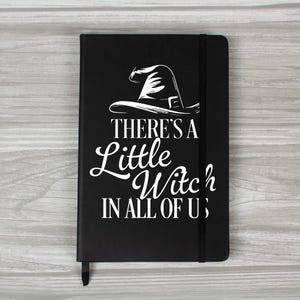 May include: Black notebook featuring the white text "THERE'S A Little Witch IN ALL OF US" and a witch hat illustration. It has a black elastic band and a ribbon bookmark. A perfect gift for Halloween.