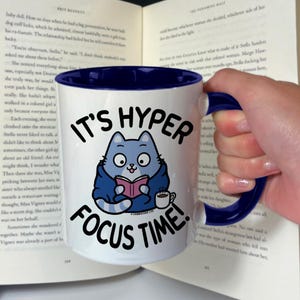 Op de afbeelding: Een witte keramische mok met een blauwe rand en handvat. De mok heeft een cartoonkat die een blauwe mantel draagt en een boek leest. De tekst "It's Hyper Focus Time!" staat op de mok.
