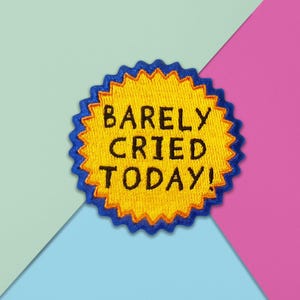 May include: Embroidered patch with a yellow centre and a blue, scalloped border. The text "BARELY CRIED TODAY!" is in black, block letters. The patch is set against a background of light blue, pink, and mint green.