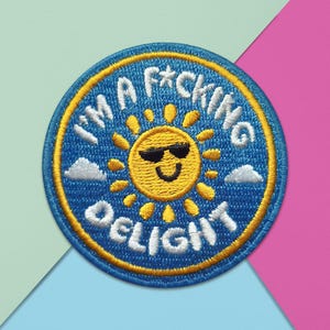 May include: A circular embroidered patch featuring a yellow sun with black sunglasses in the centre. The background is blue with small white clouds. The white text reads I'M A F*CKING DELIGHT, arched around the sun with a yellow border.