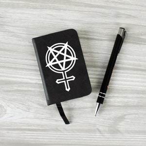 May include: A black notebook with a white pentagram and female symbol on the cover. A silver pen is next to the notebook on a wooden surface.