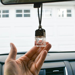 Op de afbeelding: Een heldere, vierkante auto-luchtverfrisser met een decoratieve bronzen bovenkant, hangend aan een zwart koord. De luchtverfrisser wordt in een hand gehouden, met een auto-interieur en achteruitkijkspiegel op de achtergrond.
