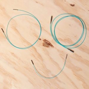 May include: Three sets of turquoise circular knitting needles with silver tips. The needles are laid out on a light wood surface.