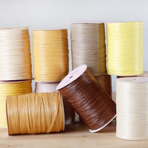 May include: Assortment of spools of colorful paper raffia ribbon in various shades of yellow, brown, and cream. The spools are arranged in a pyramid shape on a wooden surface.