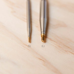 Op de afbeelding: Close-up van twee metalen breinaalden met gouden puntjes. De naalden zijn gemarkeerd met "S" en "L" voor de maat.