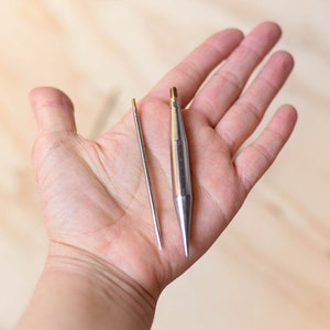 Op de afbeelding: Twee zilveren breinaalden, een lange en een korte, worden in een hand gehouden. De naalden hebben puntige punten en zijn gemaakt van metaal.