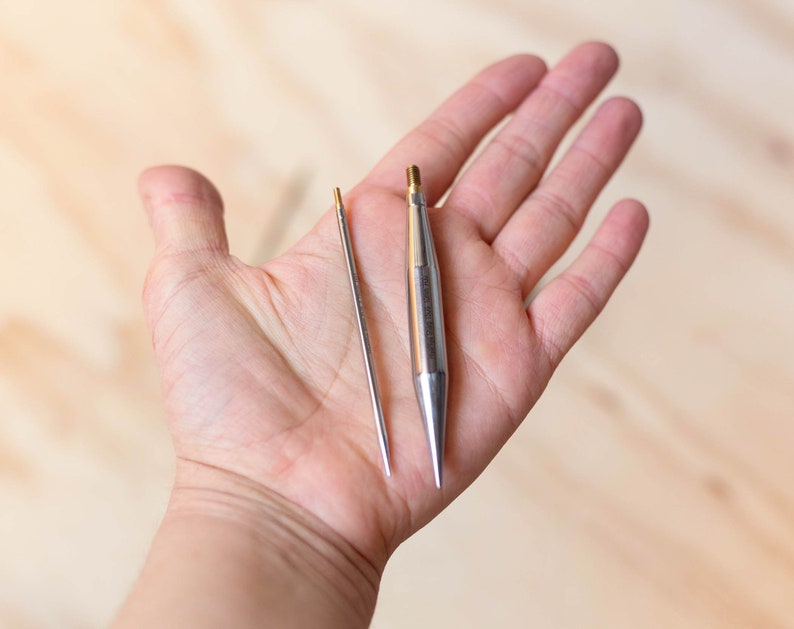 Op de afbeelding: Twee zilveren breinaalden, een lange en een korte, worden in een hand gehouden. De naalden hebben puntige punten en zijn gemaakt van metaal.