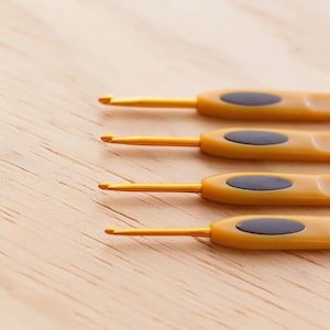 Può includere: Un set di quattro uncinetti in metallo color oro disposti in fila parallela. Ogni uncinetto presenta un manico giallo con un inserto di presa grigio scuro di forma ovale. Gli uncinetti variano nelle dimensioni e presentano marcature.