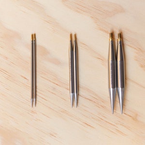 Peut inclure: Trois jeux d'aiguilles à tricoter, chacun d'une taille différente. Les aiguilles sont en métal et ont une finition argentée. Les aiguilles sont disposées en rangée sur une surface en bois brun clair.