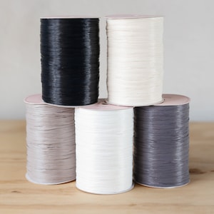 May include: Five spools of ribbon in various colors, including black, white, gray, and beige. The spools are stacked on a wooden surface.