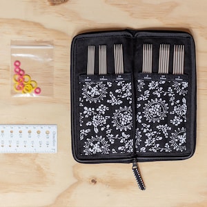 Puede incluir: Una funda negra con cremallera y un estampado floral blanco contiene un juego de 12 agujas de tejer. Las agujas est&aacute;n dispuestas en filas por tama&ntilde;o, con tama&ntilde;os de 2,25 mm, 2,5 mm, 3,0 mm, 3,25 mm, 3,5 mm y 3,75 mm. Tambi&eacute;n se muestra una bolsa de pl&aacute;stico transparente con marcadores de punto de color rosa y amarillo.