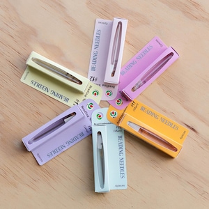May include: A collection of six colorful paper packages containing beading needles, arranged in a circular pattern. Each package features the text BEADING NEEDLES, specific size numbers like #10, #11, and #12, and indicates piece counts of 2 or 4.