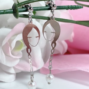 May include: A pair of silver earrings with a delicate chain and a pearl charm. Each earring features a cutout of a woman's face.