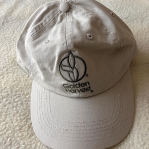 May include: A light beige baseball cap featuring a dark brown embroidered logo of a stylized plant inside a circle above the text Golden Harvest. The cap has a structured crown, a curved brim, and a fabric-covered button at the top center.