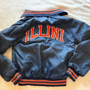 May include: A navy blue bomber jacket featuring the word ILLINI in bold orange and white lettering across the back. The jacket includes a collar, long sleeves, and ribbed cuffs and hem with orange and white stripes. The fabric has a smooth finish.
