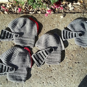 Puede incluir: Cuatro cascos de caballero de ganchillo grises con detalles en negro y rojo. Los cascos están sobre una superficie de hormigón gris.