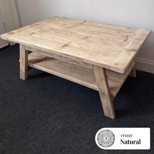 May include: A light brown, rectangular wooden coffee table with a lower shelf. The table has a rustic design with visible wood grain. The legs are angled, and the table is on a dark grey carpet. The text "FINISH Natural" is in the lower right corner. The table is made of natural wood.