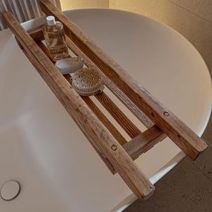 Peut inclure: Un plateau de bain en bois avec une finition en chêne naturel. Le plateau comporte trois compartiments et est rempli de produits de bain, dont une bouteille de gel douche spa, un gant de toilette et une brosse de bain.