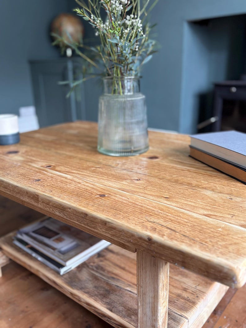 May include: A rectangular wooden coffee table with a natural light brown finish and visible wood grain, featuring a lower shelf for storage. The table surface holds a clear ribbed glass vase with green foliage and a stack of two books.