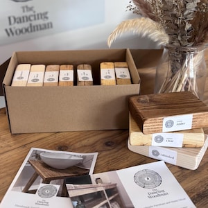 Puede incluir: Una caja de cartón llena de bloques de madera, cada uno etiquetado. Varios bloques de madera acabados están apilados cerca, etiquetados como "Ámbar", "Claro" y "Sin acabar". El logotipo de The Dancing Woodman es visible en la caja y en los materiales promocionales.