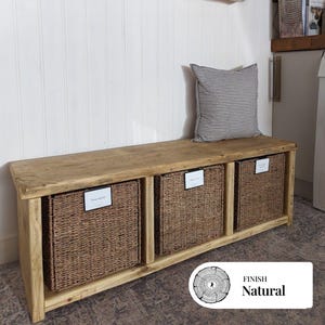 May include: A rustic wooden bench with three wicker storage baskets. The bench has a natural finish and a rectangular design. A cushion with grey stripes sits on the bench. The baskets are labelled "Trainers", "Wellies", and "Hats".