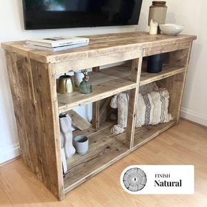 Puede incluir: Una consola de TV de madera natural con estanterías abiertas y una parte superior plana. La consola tiene un aspecto rústico de madera reciclada. Se exhiben objetos decorativos, incluidos libros, jarrones y un cuenco, en la parte superior y en los estantes. El acabado es natural.