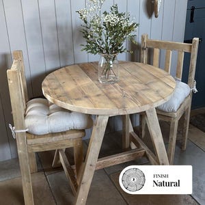 Könnte beinhalten: Ein runder Holztisch und zwei passende Stühle mit natürlicher Oberfläche. Der Tisch hat einen Durchmesser von etwa 90 cm. Die Stühle haben cremefarbene Kissen. Eine Glasvase mit weißen Blumen steht auf dem Tisch. Der Text "FINISH Natural" ist sichtbar.