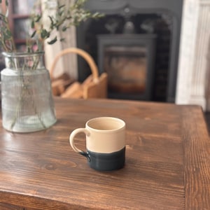 May include: A two-tone ceramic mug, cream above and dark teal below, with a rounded handle. It rests on a dark brown wooden table. The background features a fireplace and a vase holding greenery. The mug is suitable for hot beverages.