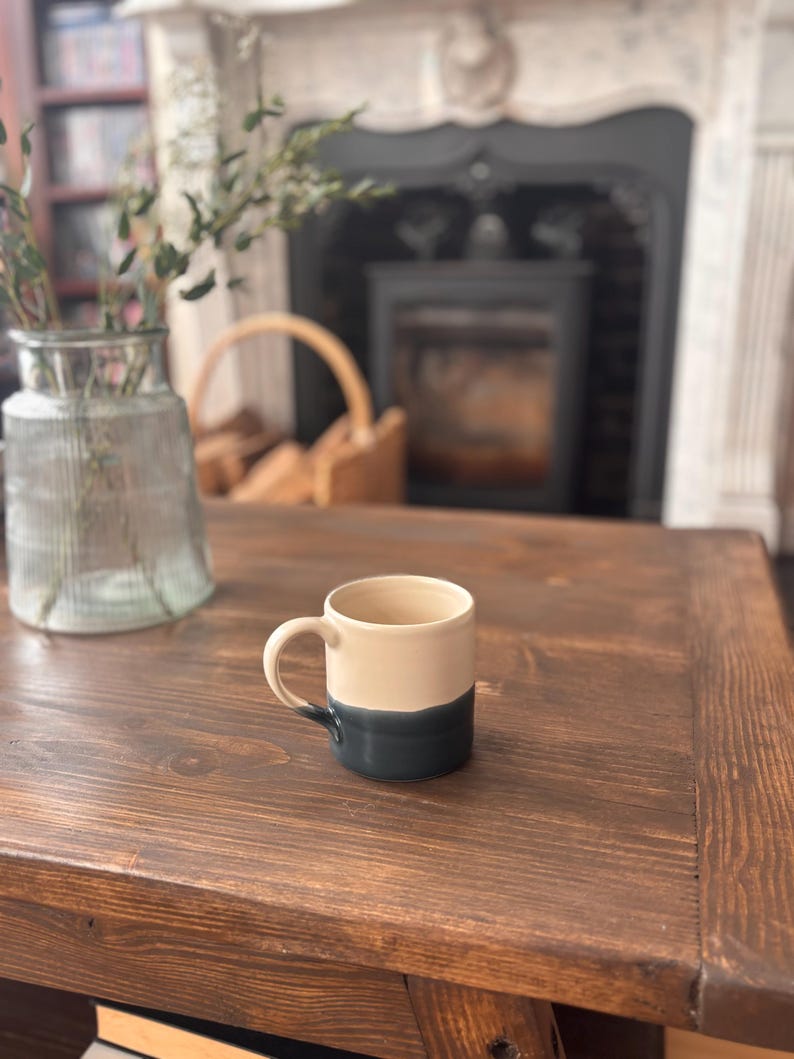 May include: A two-tone ceramic mug, cream above and dark teal below, with a rounded handle. It rests on a dark brown wooden table. The background features a fireplace and a vase holding greenery. The mug is suitable for hot beverages.