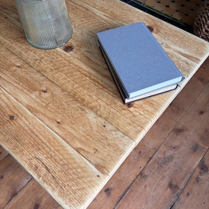 May include: A light brown wooden coffee table with a visible grain and knots, featuring a clear ribbed glass vase containing green foliage and a stack of two books with a grey cover on top, resting on a matching wooden floor surface.