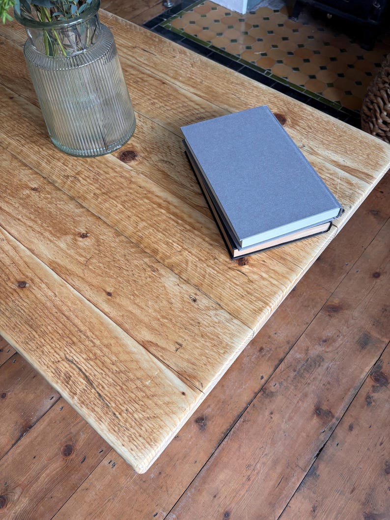 May include: A light brown wooden coffee table with a visible grain and knots, featuring a clear ribbed glass vase containing green foliage and a stack of two books with a grey cover on top, resting on a matching wooden floor surface.