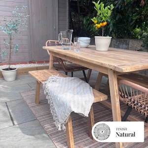 May include: A rectangular light wood dining table with a matching bench and two woven chairs on a striped outdoor rug. A potted citrus tree, a glass pitcher, and a glass tumbler sit on the table. A beige fringed throw blanket rests on the bench.