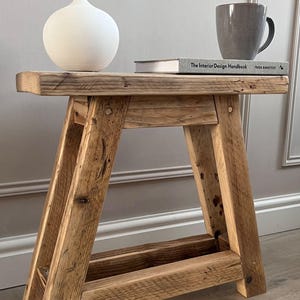 Könnte beinhalten: Rustikaler Beistelltisch aus Holz mit natürlicher Oberfläche. Der Tisch hat ein stabiles A-Rahmen-Design und ist mit einer weißen Vase, einem Buch mit dem Titel "The Interior Design Handbook" und einer grauen Tasse versehen. Der Tisch ist ca. 61 cm lang.