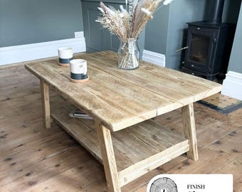 May include: A light-toned wooden coffee table with a lower shelf. The table has a clear finish and is supported by angled legs. Two mugs and a vase of dried flowers sit on top. The text "FINISH Clear" is in the bottom right corner.