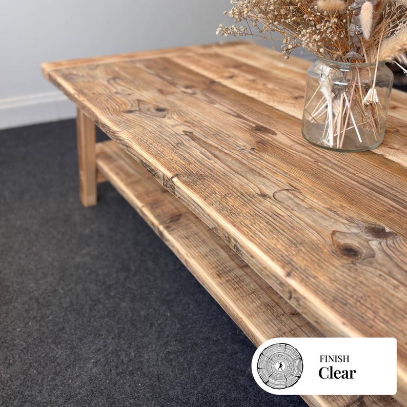May include: A wooden coffee table with a clear finish, showing the natural grain. The table has a lower shelf and is rectangular. A glass jar with dried flowers sits on the table. The table's finish is clear.