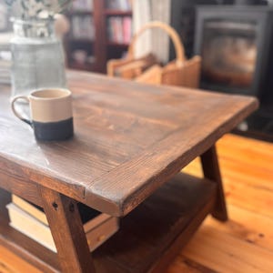 May include: A dark brown, rustic wooden coffee table with a lower shelf. A two-tone beige and navy blue mug sits on the table's surface. The table is in a living room setting with a fireplace and a vase of greenery in the background.