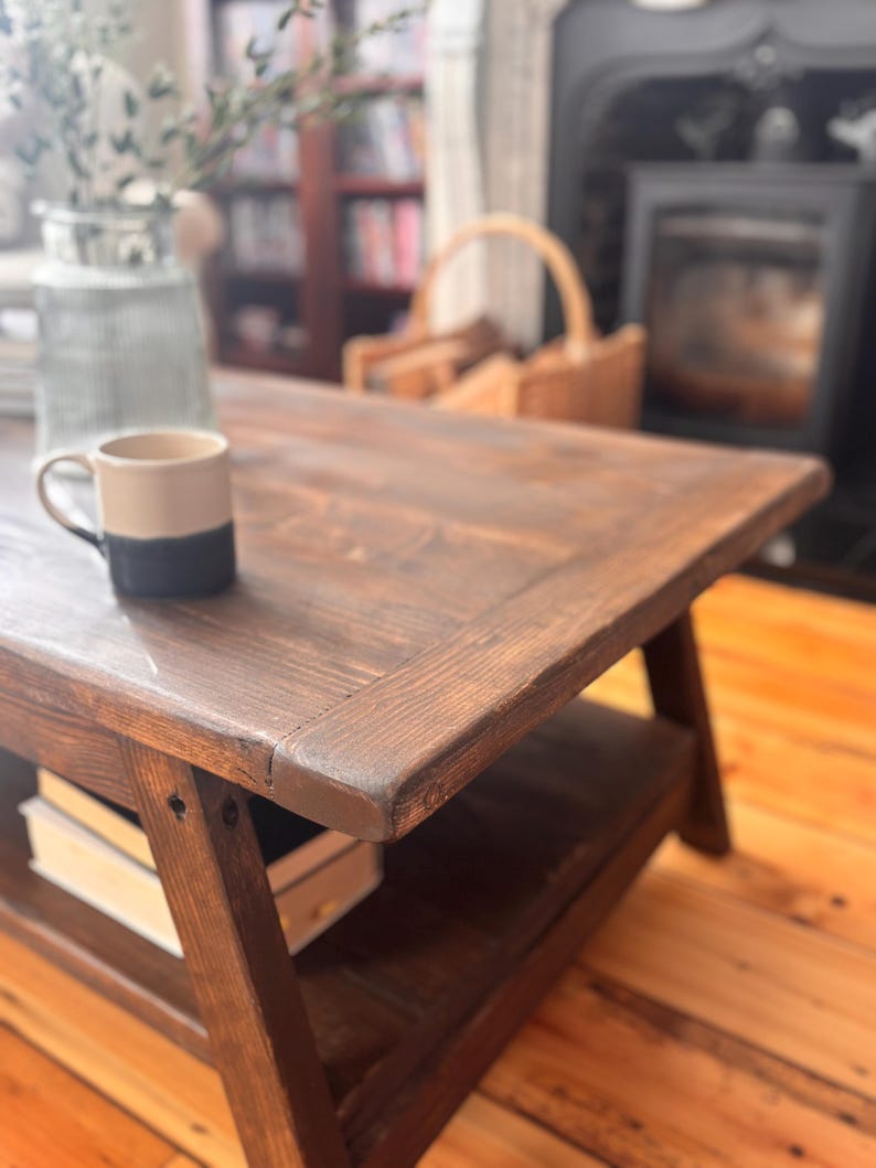 May include: A dark brown, rustic wooden coffee table with a lower shelf. A two-tone beige and navy blue mug sits on the table's surface. The table is in a living room setting with a fireplace and a vase of greenery in the background.