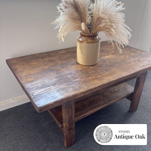 May include: A rectangular coffee table with a dark brown wood grain finish and a lower shelf. A beige ceramic vase filled with dried pampas grass sits on the surface. A small white label in the corner displays the text: FINISH Antique Oak.