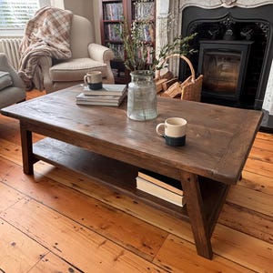 May include: A dark brown wooden coffee table with a lower shelf. Two mugs and a stack of books are on the table. The table is in a living room with a fireplace, armchair, and wooden floorboards.