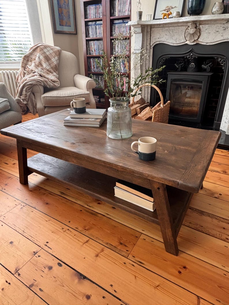 May include: A dark brown wooden coffee table with a lower shelf. Two mugs and a stack of books are on the table. The table is in a living room with a fireplace, armchair, and wooden floorboards.