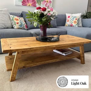 May include: A light oak rectangular coffee table with a lower shelf. The table has a natural wood grain finish. A vase of flowers sits on a decorative tray on top of the table. The table is in front of a blue sofa with floral cushions.