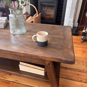 May include: A dark brown wooden coffee table with a lower shelf holding books. A two-tone mug sits on the table, and a glass vase with greenery is also present. The table is supported by angled legs.