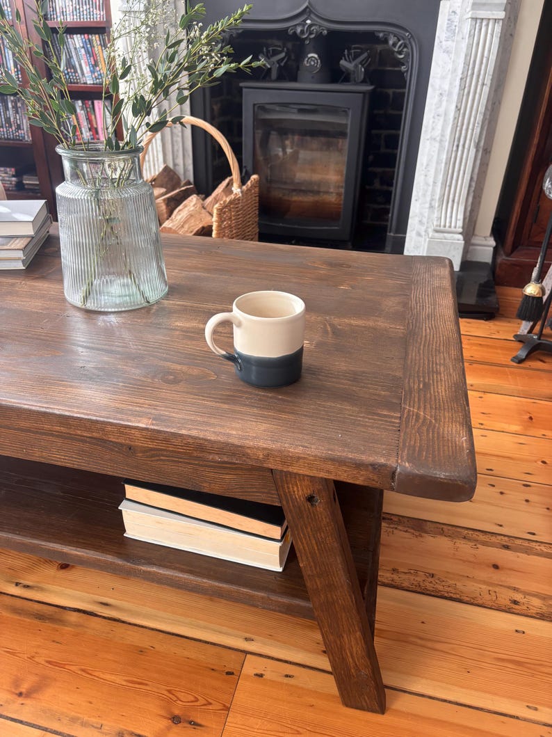 May include: A dark brown wooden coffee table with a lower shelf holding books. A two-tone mug sits on the table, and a glass vase with greenery is also present. The table is supported by angled legs.