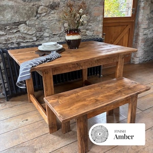 May include: A wooden dining table and bench set with an amber finish. The rectangular table has visible wood grain, and is paired with a matching bench. A vase with dried flowers and plates are on the table. The finish is labelled "Amber".