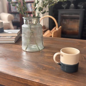 May include: A clear glass vase with vertical ridges contains green and white sprigs, on a dark brown wooden table. A cream and navy blue mug is on the table. The background features a fireplace and a bookshelf.