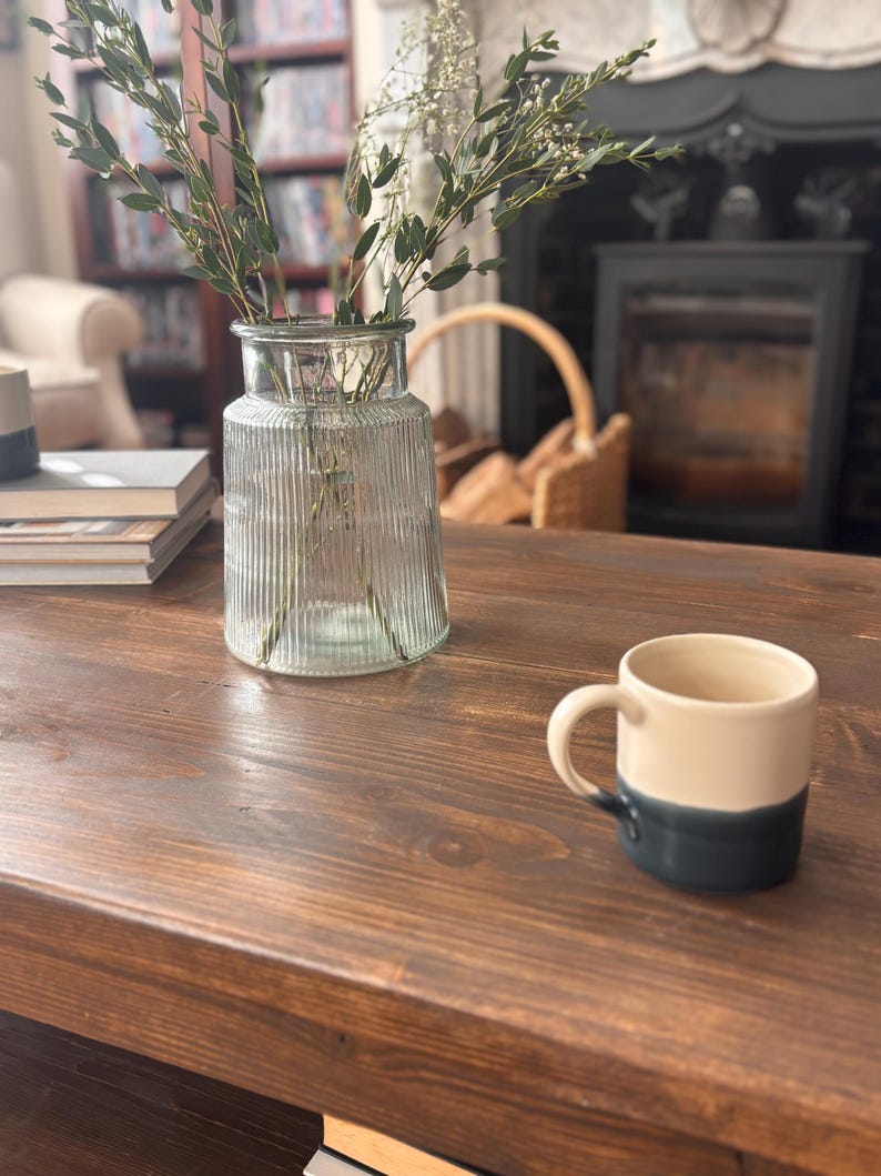 May include: A clear glass vase with vertical ridges contains green and white sprigs, on a dark brown wooden table. A cream and navy blue mug is on the table. The background features a fireplace and a bookshelf.