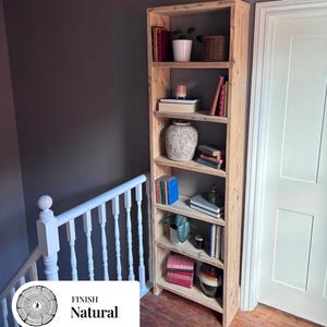 May include: A tall, narrow, light-colored wooden bookshelf with six open shelves. The shelves are filled with various items including books, a white potted plant, a woven basket, a textured ceramic vase, and a small glass bottle.
